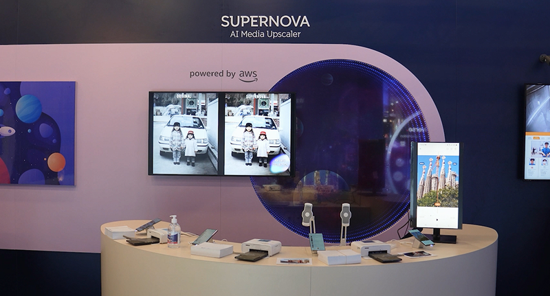 SUPERNOVA AI Media Upscaler 기술 전시 부스 전경. 중앙에는 AWS 기반 실시간 화질 개선 전후 비교 영상이 양쪽 화면에 재생되고 있으며, 부스 테이블에는 스마트폰과 디스플레이 장비들이 배치되어 있다. 벽면에는 ‘powered by AWS’와 슈퍼노바 로고가 표시되어 있다.