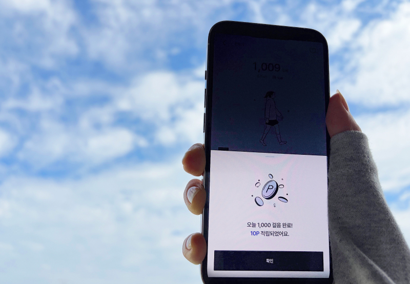 맑은 하늘을 배경으로 한 손에 스마트폰을 들고 있는 모습. 화면에는 ‘오늘 1,000걸음 완료! 10P 적립되었어요.’라는 문구가 보이며, 걷기 미션 완료 시 포인트가 적립되는 air 앱의 리워드 화면을 보여줍니다.