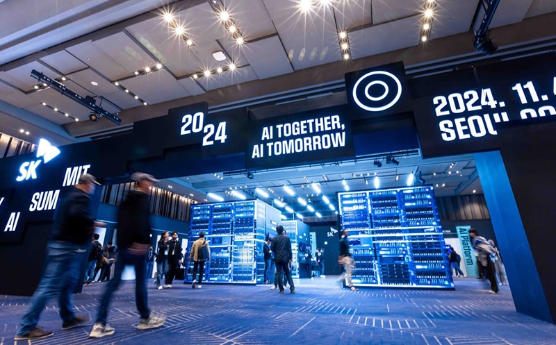 2024 SK AI SUMMIT 행사장 입구 전경. 천장에 조명이 밝게 비추는 전시장 내부에서 사람들이 이동하고 있으며, 입구 상단에는 “2024”, “AI TOGETHER, AI TOMORROW”, “2024. 11. SEOUL COEX” 문구가 밝은 흰색으로 표시되어 있음. 