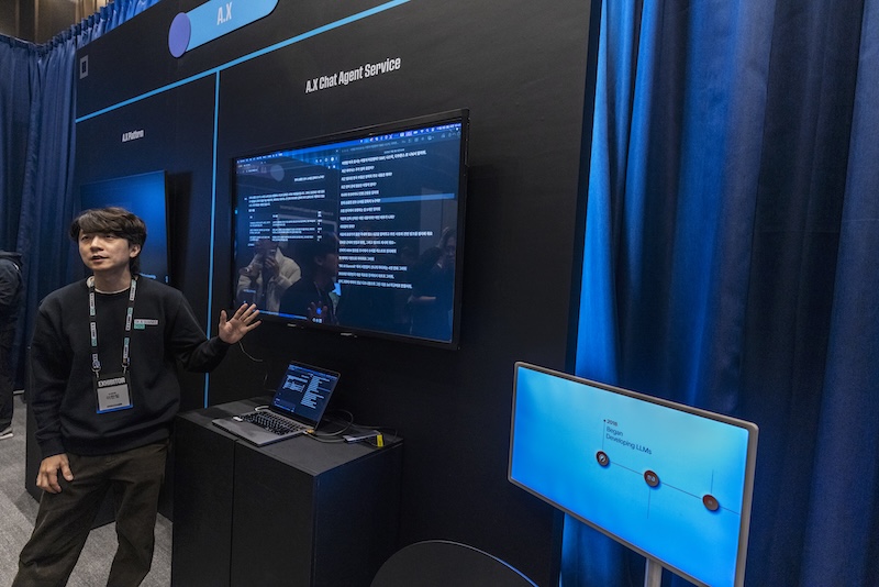 ‘A.X Chat Agent Services’ 부스에서 담당자가 AI 에이전트 대시보드를 설명 중이다. 실시간 대화 로그와 에이전트 선택 UI가 모니터에 보인다.