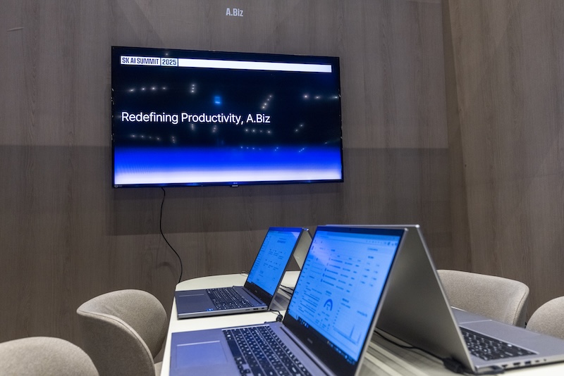 ‘Redefining Productivity, A.Biz’ 문구 아래 원형 테이블 위에 노트북들이 놓여 있다. 각 화면엔 A.Biz의 협업 인터페이스가 실행 중이다.