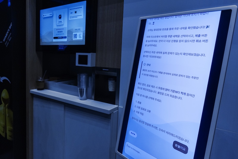 오른쪽 대형 스크린에는 자판기 고객 불만을 처리하는 AI 챗봇 UI가, 왼쪽 자판기엔 음료 메뉴 화면이 보인다. 자판기 운영 전 과정을 자동화한 모습이다.