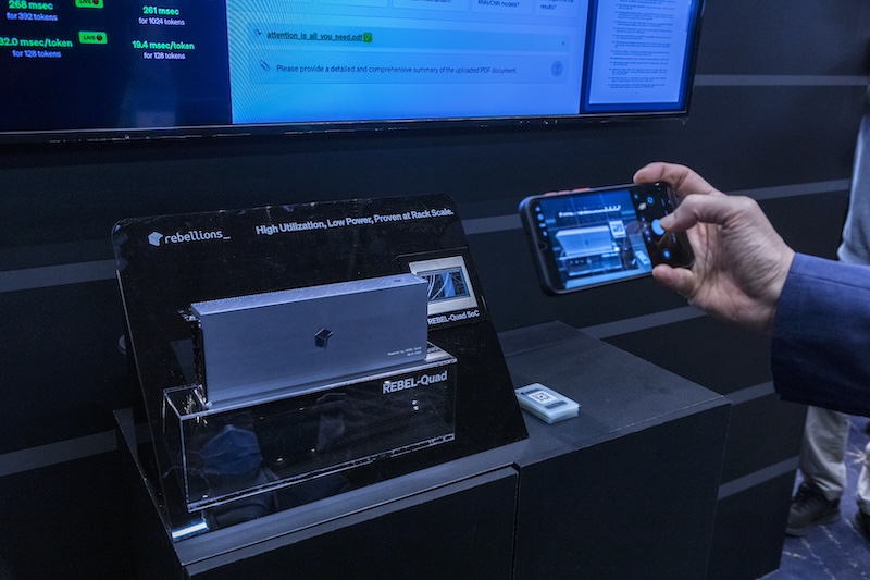 리벨리온스 부스의 REBEL-Quad 칩셋이 투명 케이스에 전시되어 있다. 한 관람객이 스마트폰으로 제품을 촬영하고 있다.