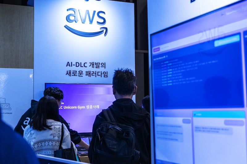 AWS 부스에서 ‘AI-DLC 개발의 새로운 패러다임’ 주제로 참가자들이 시연을 관람하고 있다. 화면에는 Unicorn Gym 성공사례가 소개되고 있다.