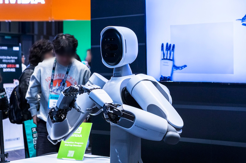 전시장에 전시된 리얼월드의 흰색 휴머노이드 로봇이 팔을 움직이며 시연 중이다. 뒤편에는 관람객과 로봇 손 모형이 함께 보인다.