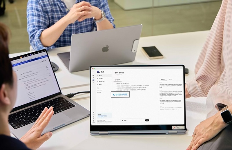 회의실 테이블에 노트북 3대를 두고 사람들이 협업하는 모습. 중앙에는 태블릿 형태의 노트북이 놓여 있으며 화면에는 에이닷 노트가 실행되고 있다. 실시간 회의 요약 기능이 표시된 노트 서비스 인터페이스가 보인다. 왼쪽 사람은 노트북으로 문서를 확인 중이고, 오른쪽 사람은 스마트폰을 옆에 둔 채 화면을 바라보고 있다. 테이블 위에는 펜과 스마트폰 등이 놓여 있으며, 실무형 회의 분위기가 연출되어 있다.