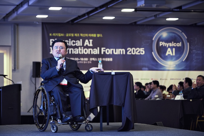 Physical AI International Forum 2025 행사 무대에서 박태완 정책관이 휠체어에 앉아 발표를 진행하고 있다. 배경에는 행사명이 적힌 대형 현수막이 걸려 있다.