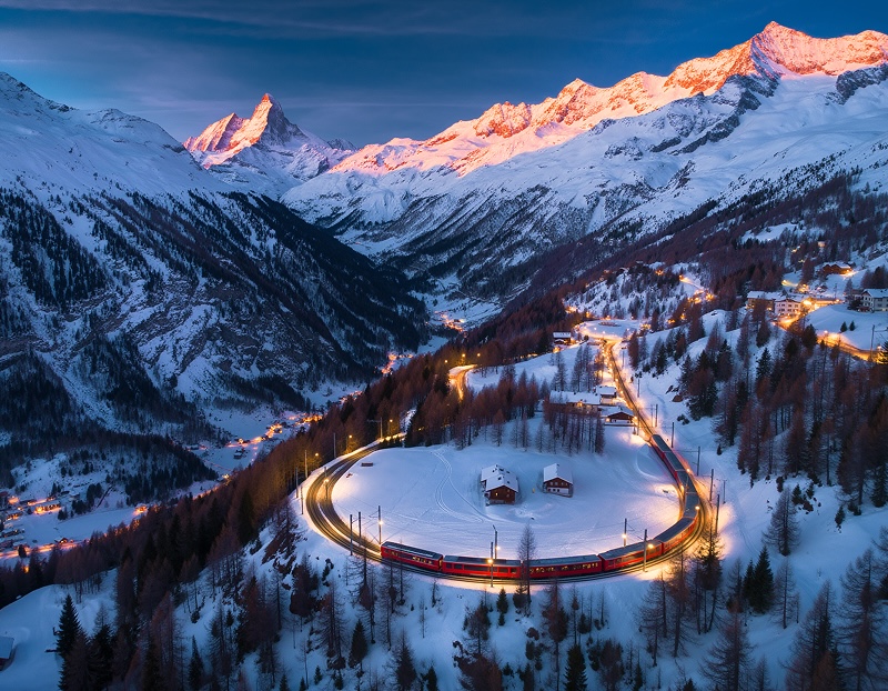 설산 아래 굽이진 철로를 따라 붉은 기차가 달리고 있다. 해 질 무렵 산 정상은 붉게 물들고 마을 불빛이 켜져 있다.