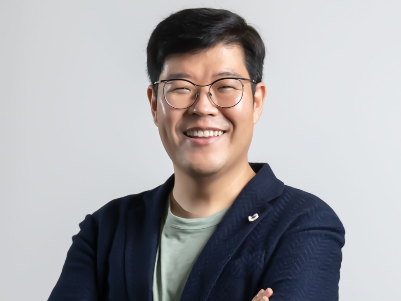 리얼월드 류중희 대표가 팔짱을 낀 채 미소 짓고 있다. 회색 배경의 프로필 사진.