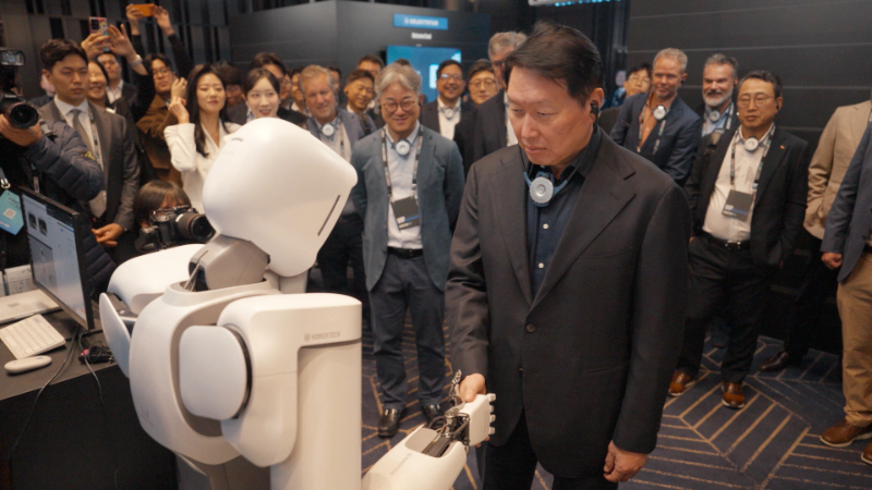 전시장 안에서 ALLEX 로봇이 최태원 회장과 악수하고 있다. 주위 관람객들이 둘러서서 지켜보고 있다.