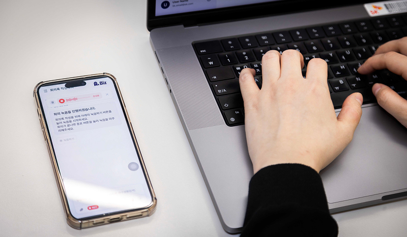 노트북에 키보드에 손을 올린 사람과 좌측에 놓인 스마트폰. 스마트폰에는 에이닷 비즈 앱이 구동되고 있다. '회의 녹음을 진행하겠습니다'라는 문구와 함께 녹음을 진행 중이다.
