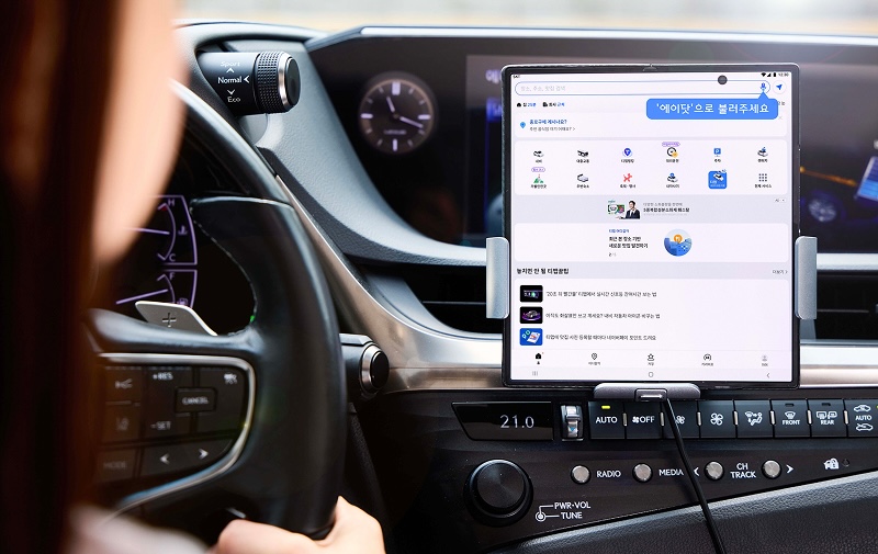 자동차 운전석에서 운전자가 대시보드 중앙에 거치된 태블릿 화면을 바라보고 있다. 태블릿에는 음성 명령 입력창과 '에이닷' 서비스 화면이 펼쳐져 있으며, 여러 아이콘과 추천 콘텐츠가 보인다.