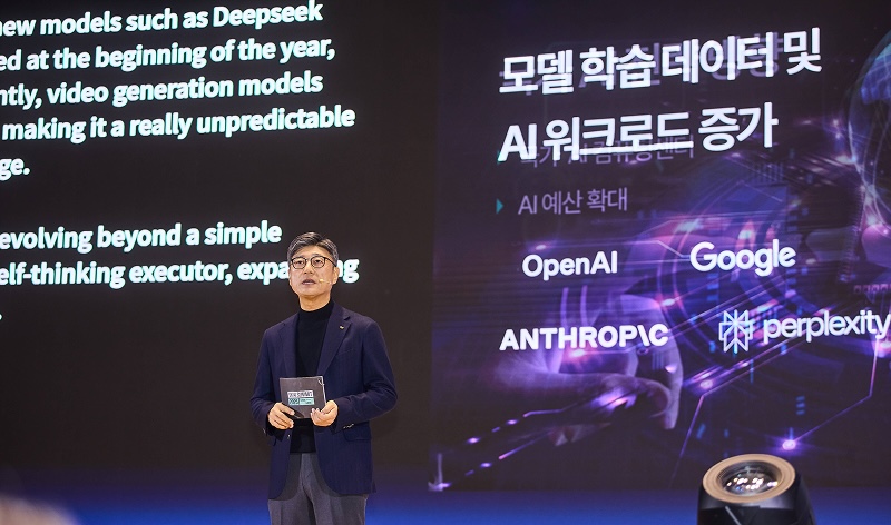 SK AI Summit 2025 무대 위 정재헌 SKT CEO. 대형 스크린에는 '모델 학습데이터 및 AI 워크로드 증가'라는 제목과 함께 OpenAI, Google, ANTHROPIC, perplexity 등의 로고가 배치되어 있다