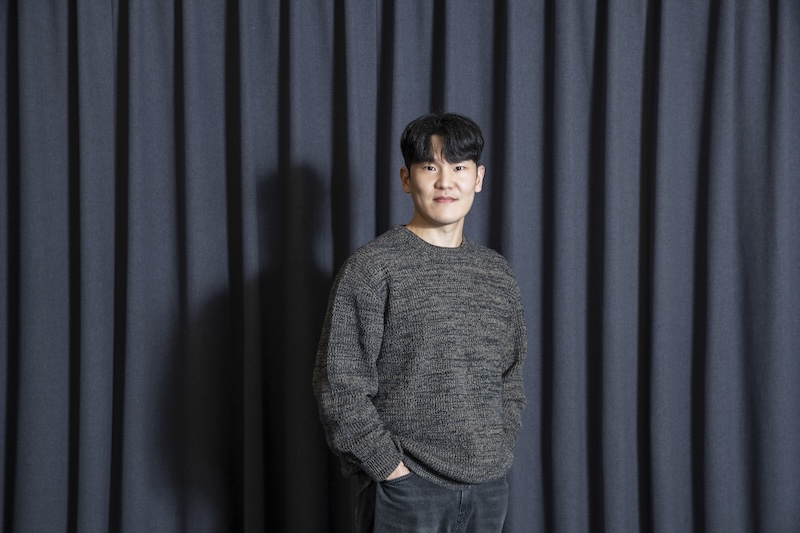 어두운 커튼 앞에서 이경필 님이 한 손을 주머니에 넣고 서서 카메라를 바라보는 인물 사진.