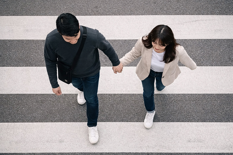 횡단보도를 위에서 내려다본 구도로 두 사람이 손을 잡고 걷고 있는 AI 이미지. 왼쪽 인물은 어두운 상의와 청바지, 흰 운동화를 착용하고 크로스백을 메고 있으며, 오른쪽 인물은 밝은 재킷과 흰 상의, 청바지, 흰 운동화를 착용하고 있다. 바닥에는 흰색과 회색의 횡단보도 줄무늬가 보인다.