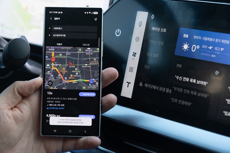 차량 내부에서 스마트폰과 차량 디스플레이가 함께 보이는 장면. 왼쪽에는 손에 들린 스마트폰 화면에 지도 내비게이션이 실행되어 있으며, 출발지는 서울특별시, 도착지는 남산서울타워로 설정되어 있다. 지도 화면에는 도로 상황과 경로가 표시되어 있고, '내 차로 목적지를 보냈어요. 자동차 내비게이션에서 확인해 주세요' 버튼 문구가 나와 있다. 오른쪽 차량 디스플레이에는 에이닷 오토 인터페이스가 표시되어 있으며, 에이닷에서 보낸 장소 문구가 표시되어 있따.