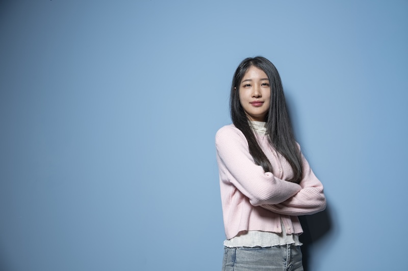 파란 배경 앞에서 팔짱을 낀 이유진 님. 분홍색 가디건을 입고 정면을 바라보고 있다.