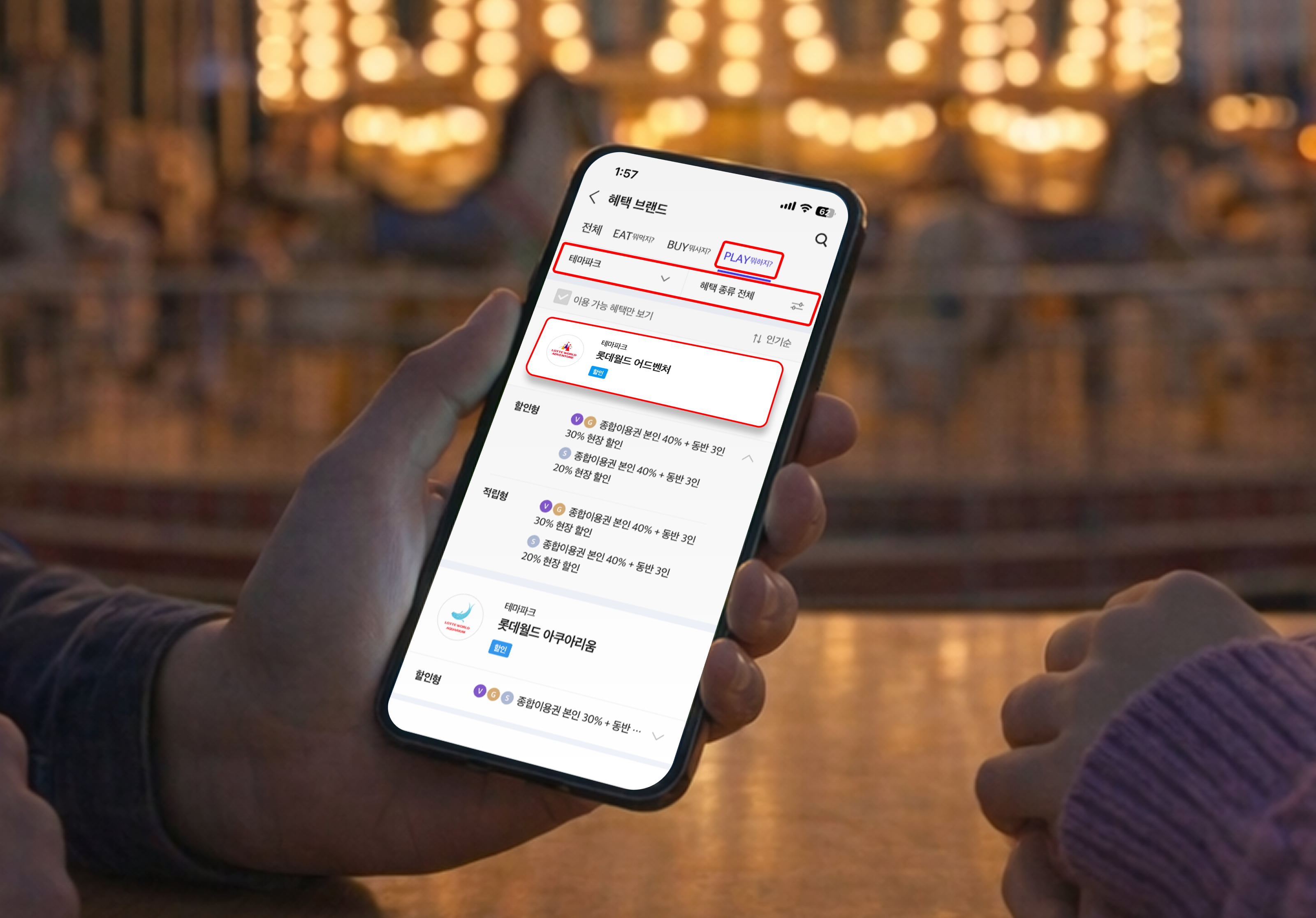 놀이공원 회전목마 조명이 흐릿하게 보이는 배경 앞에서 스마트폰을 들고 있는 장면. 스마트폰 화면에는 ‘혜택 브랜드’ 메뉴와 함께 PLAY 영역이 선택되어 있으며 ‘롯데월드 어드벤처’ 할인 혜택 정보가 강조 표시되어 있어 T멤버십으로 놀이공원 혜택을 확인하는 모습을 보여준다.