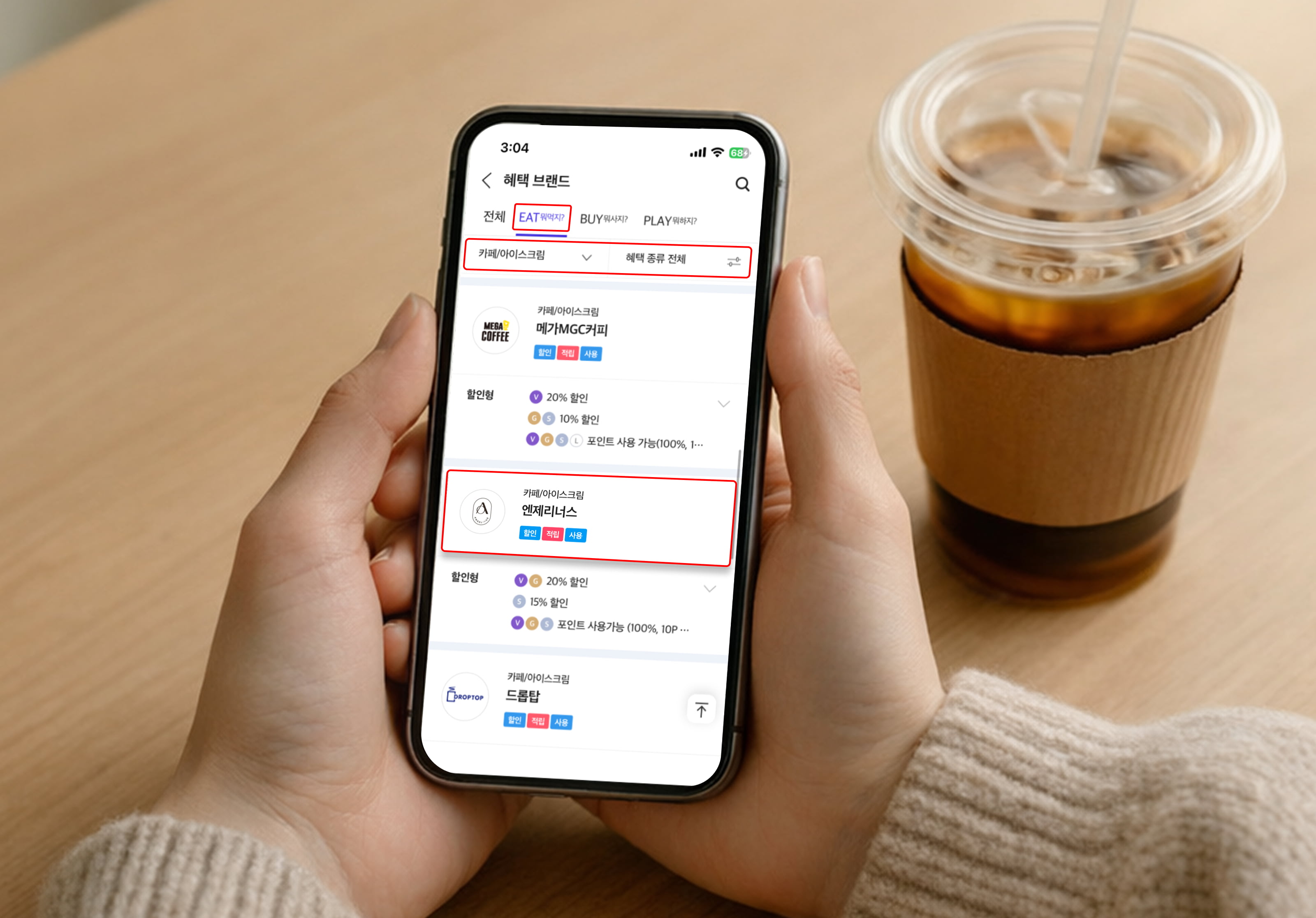 테이블 위 아이스커피 컵 옆에서 스마트폰을 양손으로 들고 있는 장면. 스마트폰 화면에는 ‘혜택 브랜드’ 메뉴의 EAT 영역이 선택되어 있으며 ‘메가MGC커피’, ‘엔제리너스’ 등 카페 브랜드와 할인 혜택 정보가 표시되어 있어 T멤버십으로 카페 할인 혜택을 확인하는 화면을 보여준다.