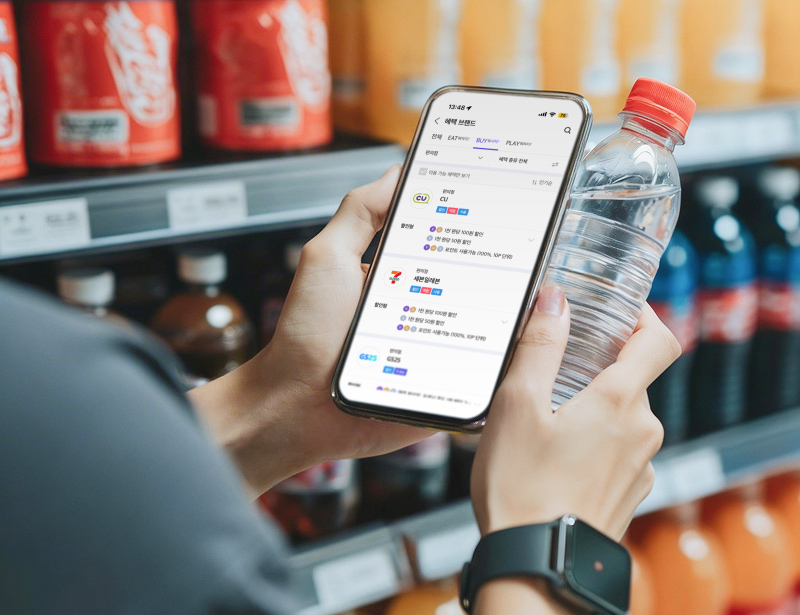 편의점 음료 진열대 앞에서 물을 들고 스마트폰의 T멤버십 혜택 화면을 확인하는 모습