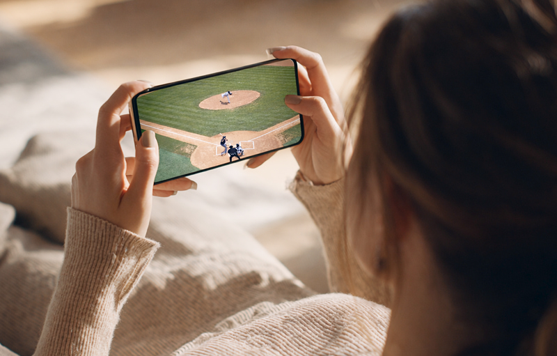 집에서 소파에 앉아 스마트폰으로 야구 경기를 시청하는 모습, 화면에는 투수와 타자가 있는 야구 경기 장면이 보이는 이미지