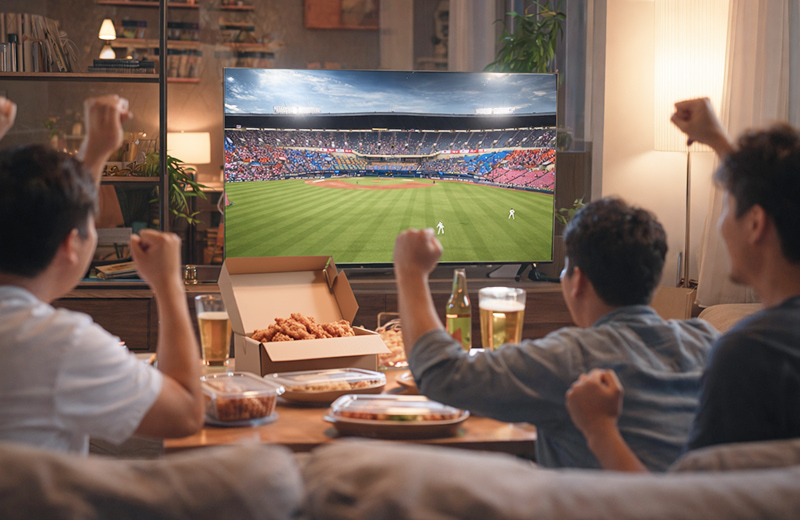 거실에서 TV로 야구 경기를 시청하며 치킨과 맥주를 즐기는 남성들이 함께 응원하는 모습, 화면에는 야구장 전경과 경기 장면이 보이는 이미지