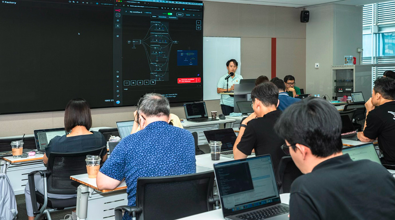 강의실에서 발표자가 마이크를 들고 AI 워크플로우 화면이 표시된 대형 스크린 앞에서 설명하고 있으며, SKT 구성원들이 노트북을 사용하며 ‘AI 프런티어’ 프로그램 교육에 참여해 실무에 적용할 AI 역량을 학습하는 모습.